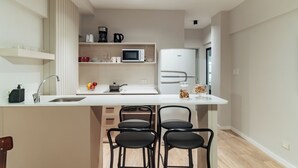 A modern kitchen with a white countertop, a built-in microwave, a refrigerator, and a sink. There are black chairs and a small island with a sink.