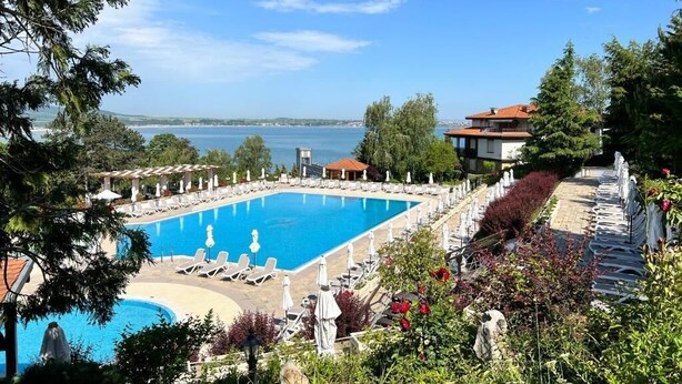 A large rectangular pool with lounge chairs and umbrellas, surrounded by greenery and a view of the sea.