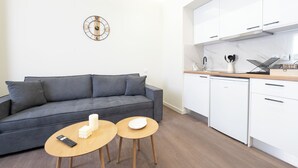 A modern living room with a grey sofa, two wooden side tables, a wall clock, and a kitchen area with white cabinets and a dishwasher. - Modern Room Exarchia Room 4 (Αθήνα)