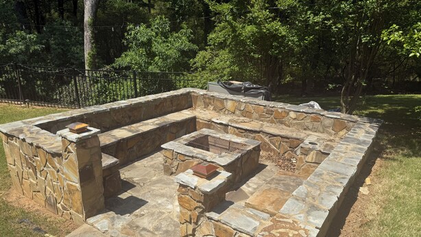 A stone-built outdoor kitchen area with a grill and a fire pit, surrounded by a stone wall and a grassy area. - FIFA Ready! Spacious 1800 sqf (Ellenwood)