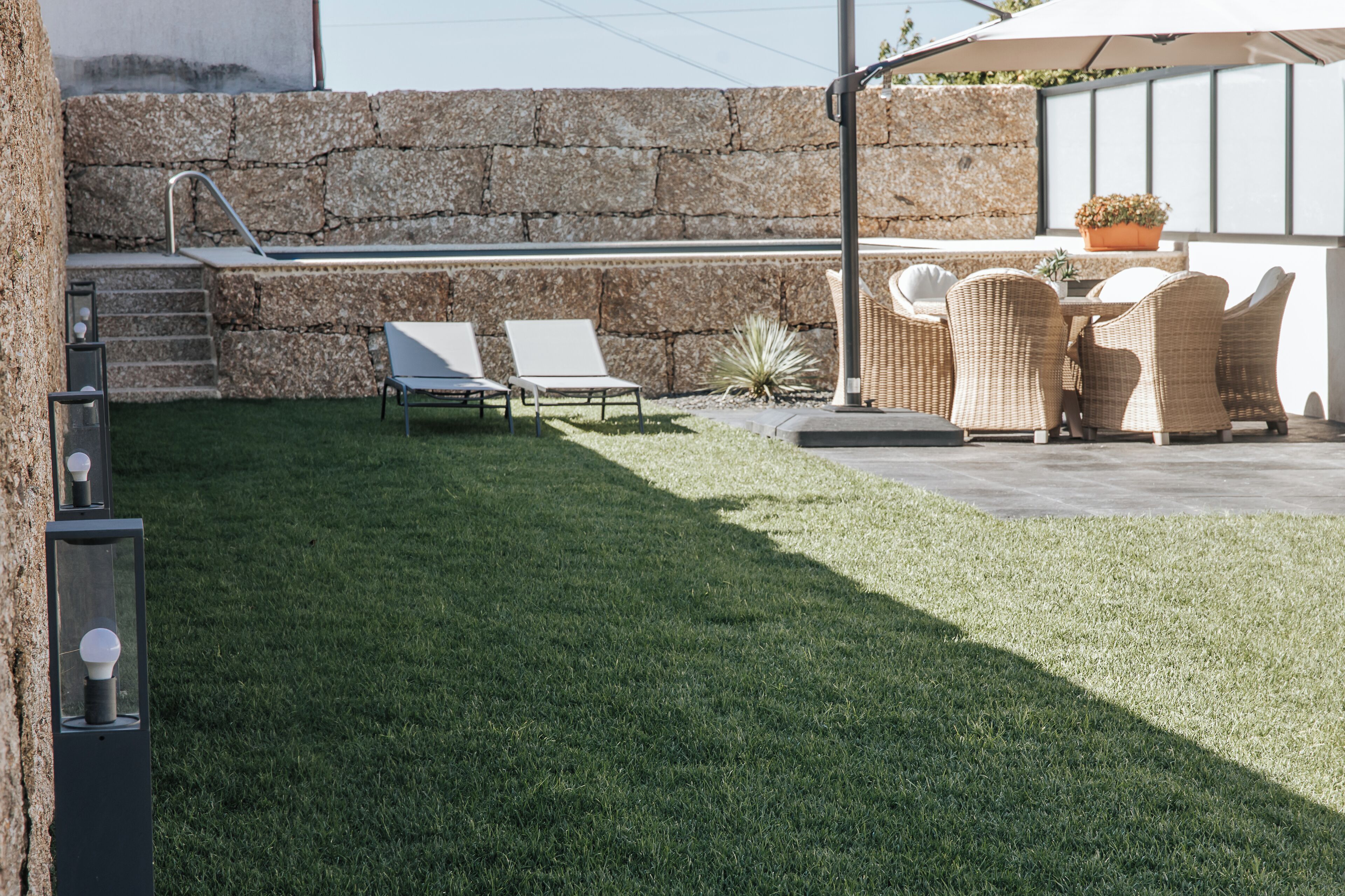 A backyard with a stone wall, a grill, two lounge chairs, a dining table with wicker chairs, and a potted plant.