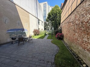 Une cour avec un chemin pavé, un espace repas comprenant des chaises et une table, un mur en briques et une zone herbeuse agrémentée d’arbustes et de fleurs.