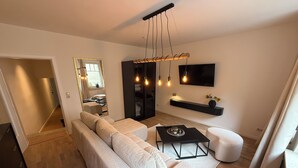 A modern living room with a sectional sofa, a coffee table, a TV mounted on the wall, and a hanging light fixture. - Stylish apartment right next to the market and town hall (Stralsund)
