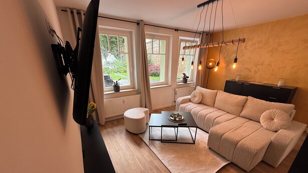 A modern living room with a sofa, a coffee table, a TV mounted on the wall, and a window with curtains. - Stylish apartment right next to the market and town hall (Stralsund)