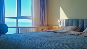 A hotel room with a large window, a bed with a blue quilt, and a view of the sea. - Chic 1-bedroom apartment in charming Batumi with AC, WiFi (Batumi)