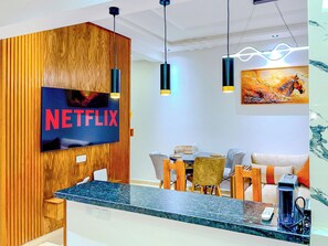 A modern interior with a wooden reception desk, a wall-mounted TV displaying 'NETFLIX,' a dining area with a table and chairs, and a ceiling with pendant lights.