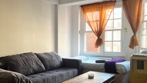 A living room with a grey sofa, a wooden coffee table, and a radiator. There is a window with orange curtains and a view of a brick building. - Marble Arch - Central London Apartment - Air Conditioner (London)