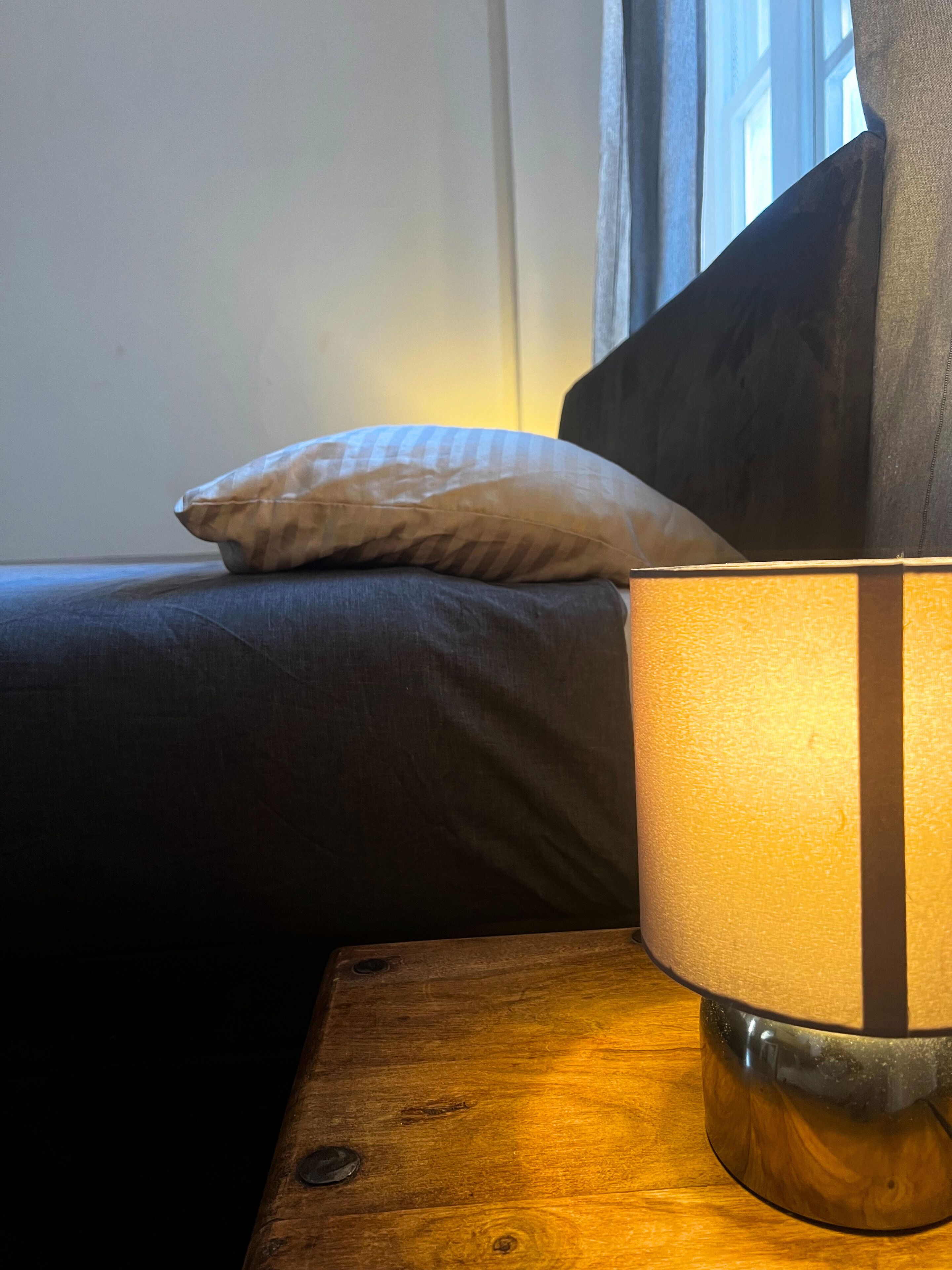 A lamp on a wooden nightstand next to a bed with a pillow.