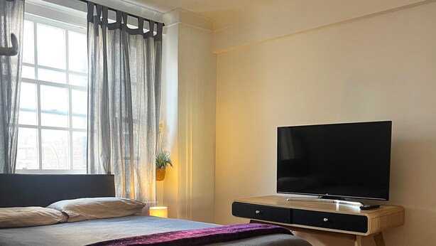 A bedroom with a bed, a TV on a wooden stand, a window with curtains, and a plant. - Marble Arch - Central London Apartment - Air Conditioner (London)