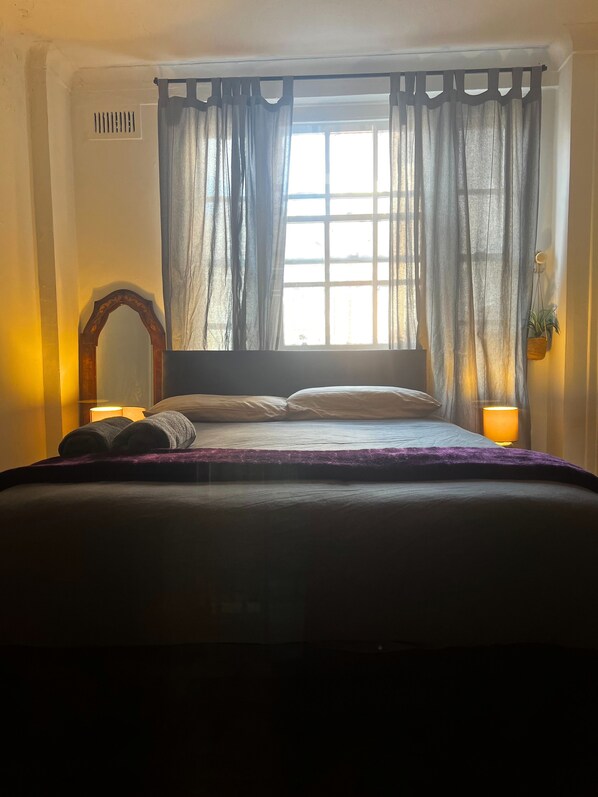 A bedroom with a bed, two bedside lamps, a window with curtains, a wall-mounted mirror, and a small plant.