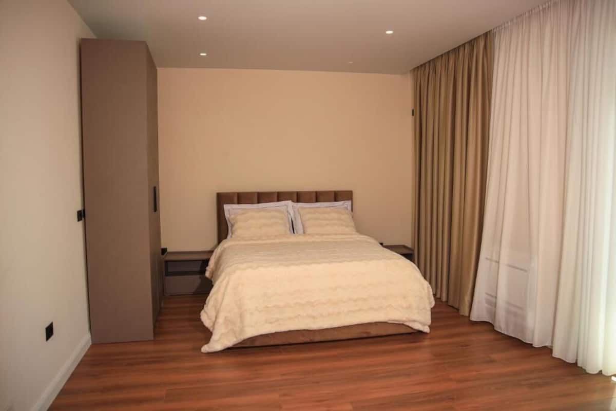 A bedroom with a wooden floor, a bed with a beige duvet, and a large window with curtains.