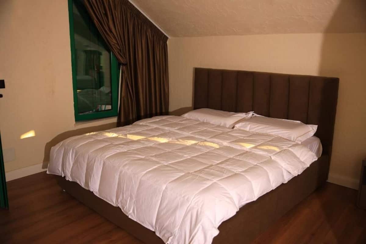 A neatly made bed with a white duvet and brown headboard in a room with wooden flooring and a window with curtains.