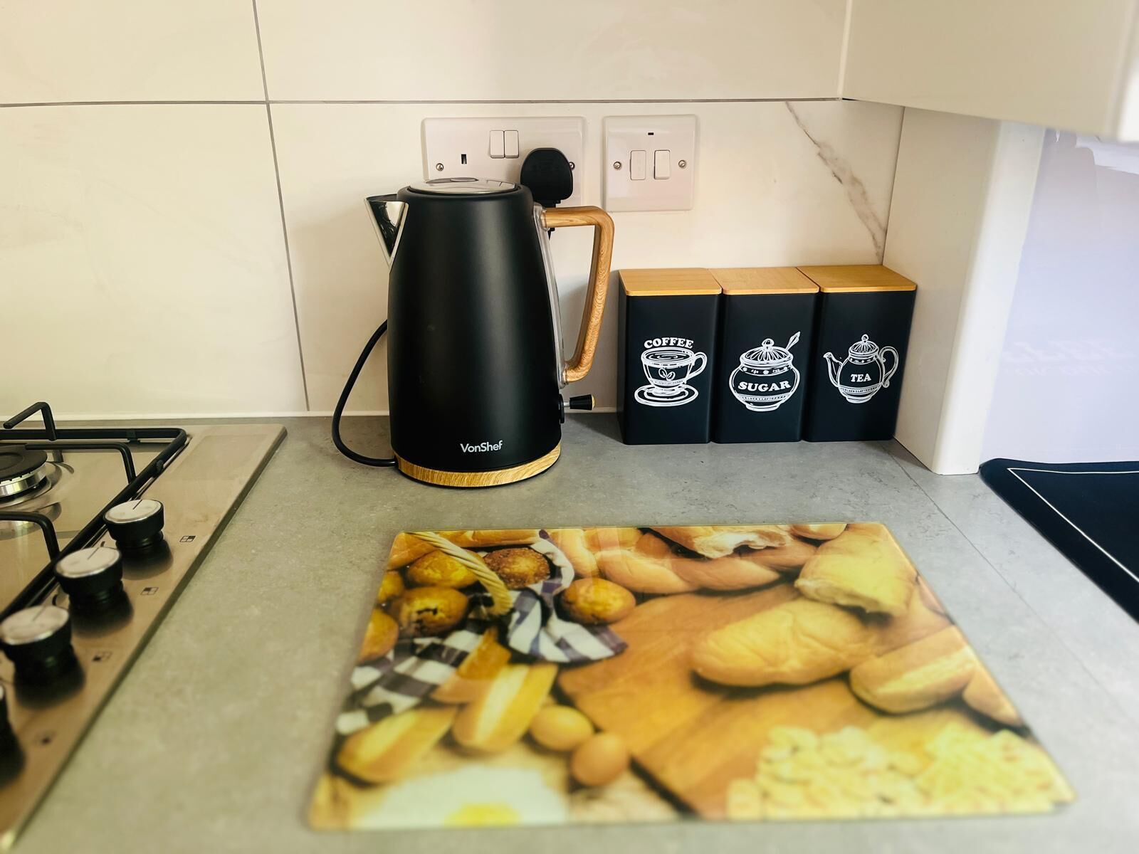 A kitchen with a gas stove, a black coffee maker, and three black boxes with white illustrations of coffee, sugar, and tea.