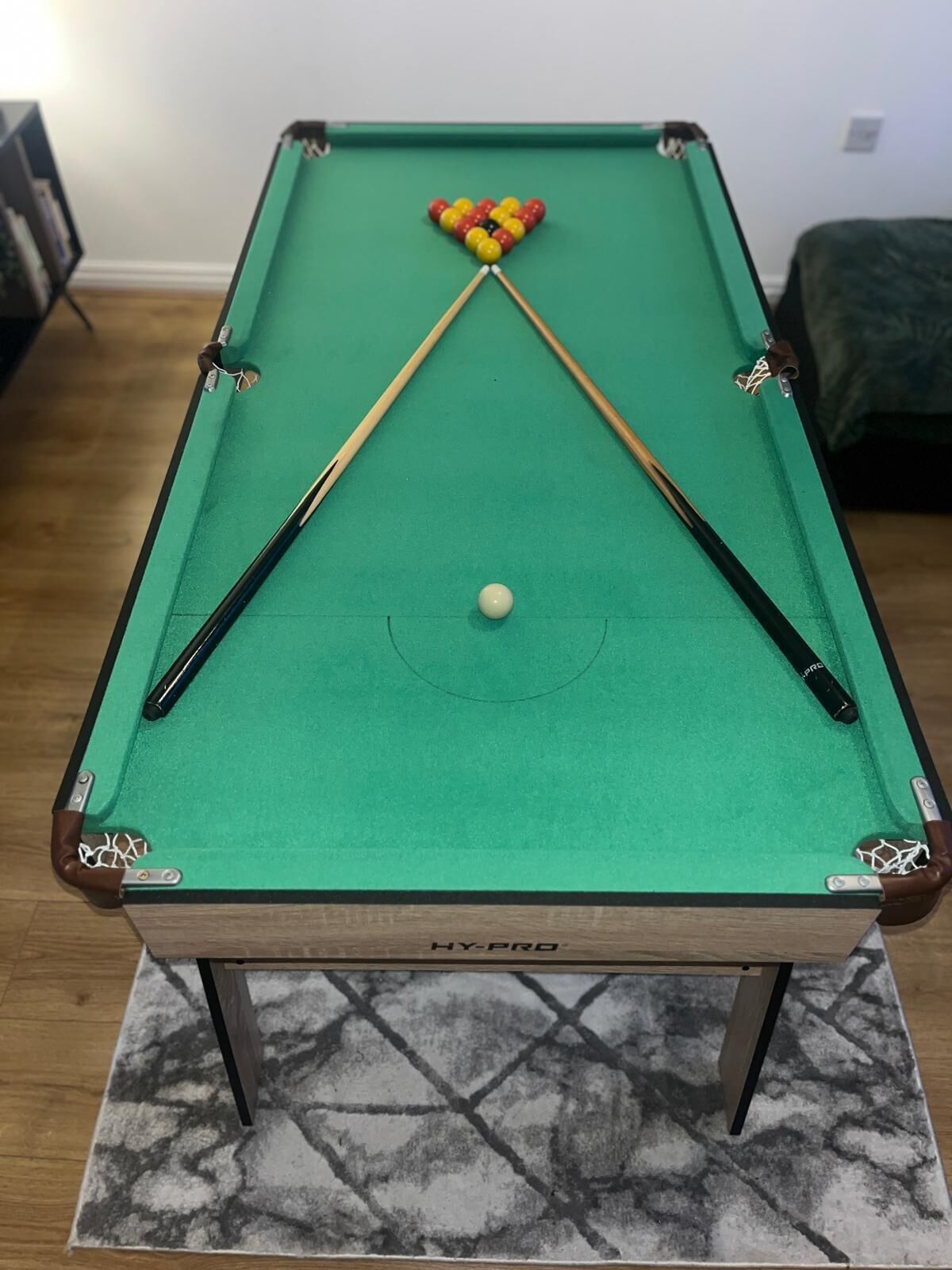 Uma mesa de bilhar com duas baquetas e uma bola branca, superfície em feltro verde, e o logotipo "HY-PRO" na lateral da mesa.