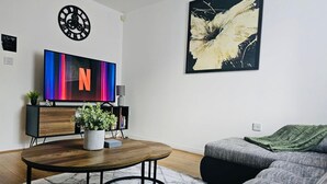 Una sala moderna con una mesa de centro de madera, un sofá gris, un televisor con una 'N' colorida en la pantalla, un reloj y arte abstracto en la pared.