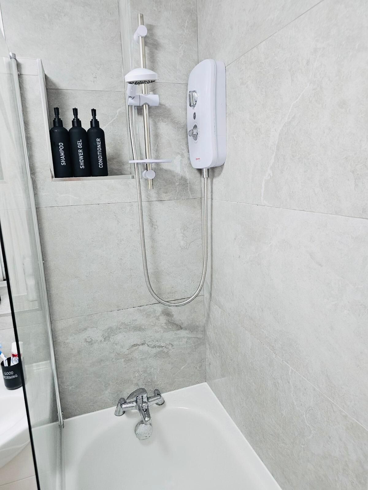 A bathroom with a bathtub, a shower, and a shelf with three bottles labeled shampoo, shower gel, and conditioner.