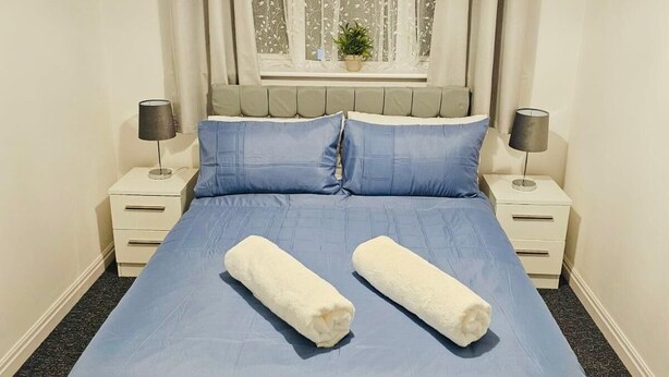 Una cama bien hecha con ropa de cama azul y dos toallas enrolladas, flanqueada por mesitas de noche blancas con lámparas, una ventana con cortinas traslúcidas y un candelabro arriba.