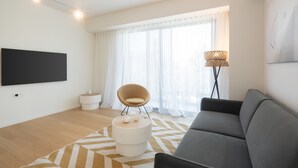 A modern living room with a grey sofa, a round white ottoman, a beige chair, a flat-screen TV mounted on the wall, and a floor lamp. - LIV Athinum 1 - Luxury Apartment 2A (Athens)