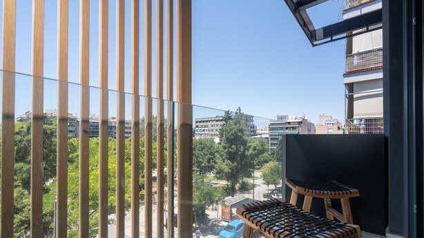 A balcony with a view of a cityscape, a wooden chair, and a small table. - LIV Athinum 1 - Luxury Apartment 2A (Athens)