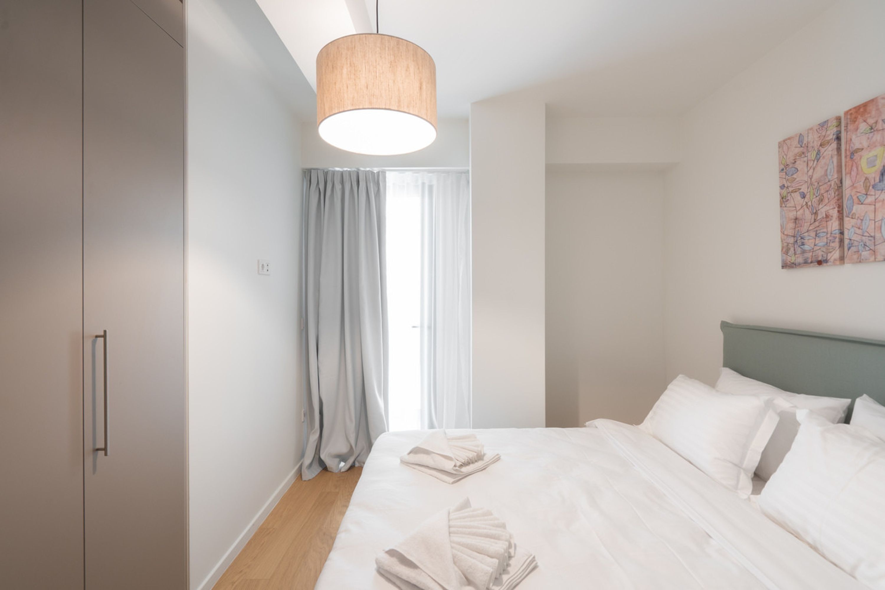 A bedroom with a bed, a hanging light fixture, a window with curtains, and a wall with artwork.
