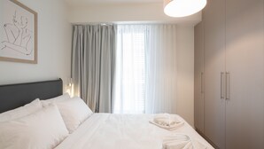 A hotel room with a bed, a wardrobe, a window with curtains, and a wall-mounted light fixture. - LIV Athinum 1 - Luxury Apartment 2A (Athens)