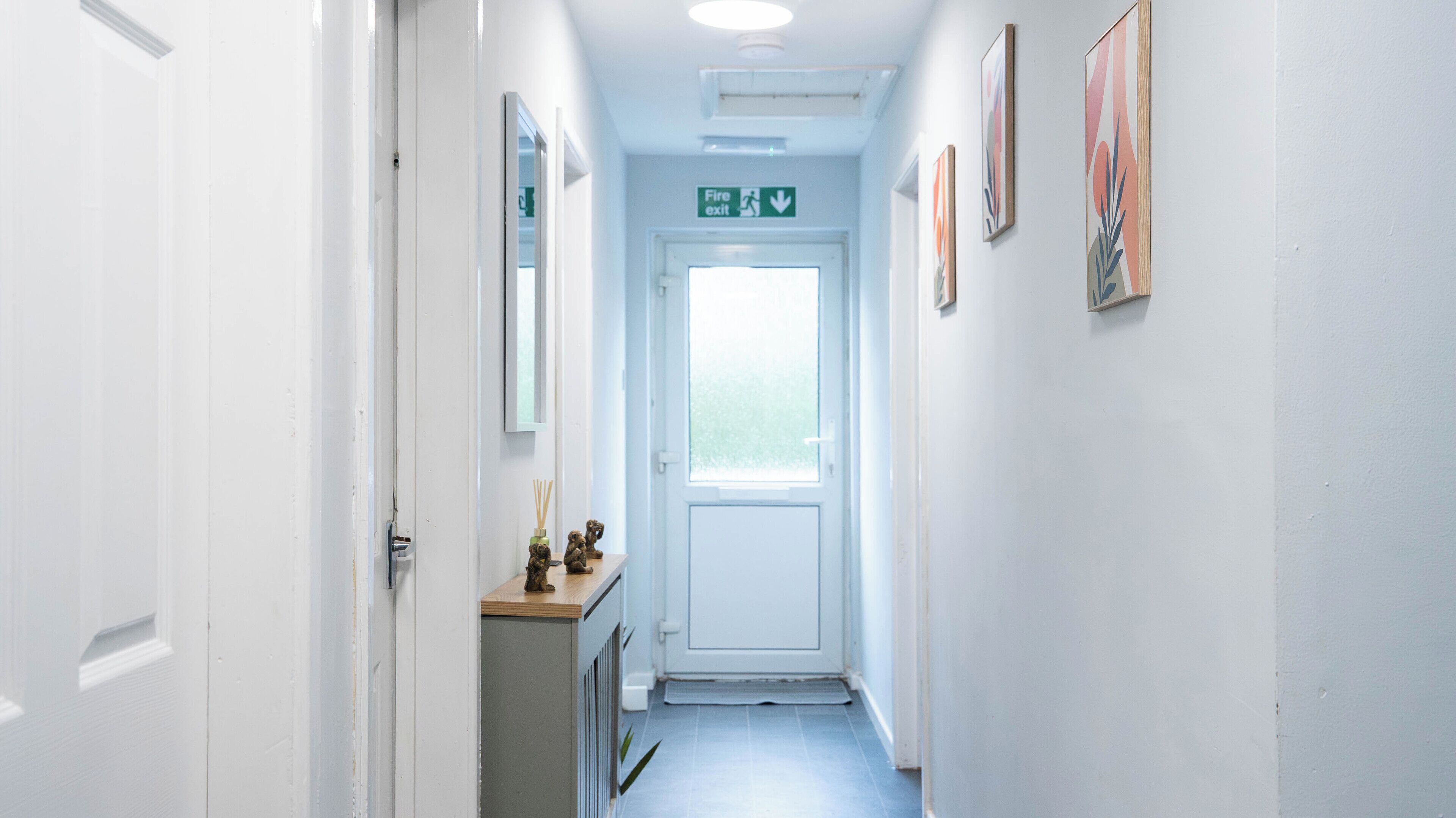 Un couloir étroit, avec une porte blanche au fond, un panneau indiquant la sortie de secours au-dessus, et une petite armoire sur le côté gauche, sur laquelle est posé un objet décoratif.
