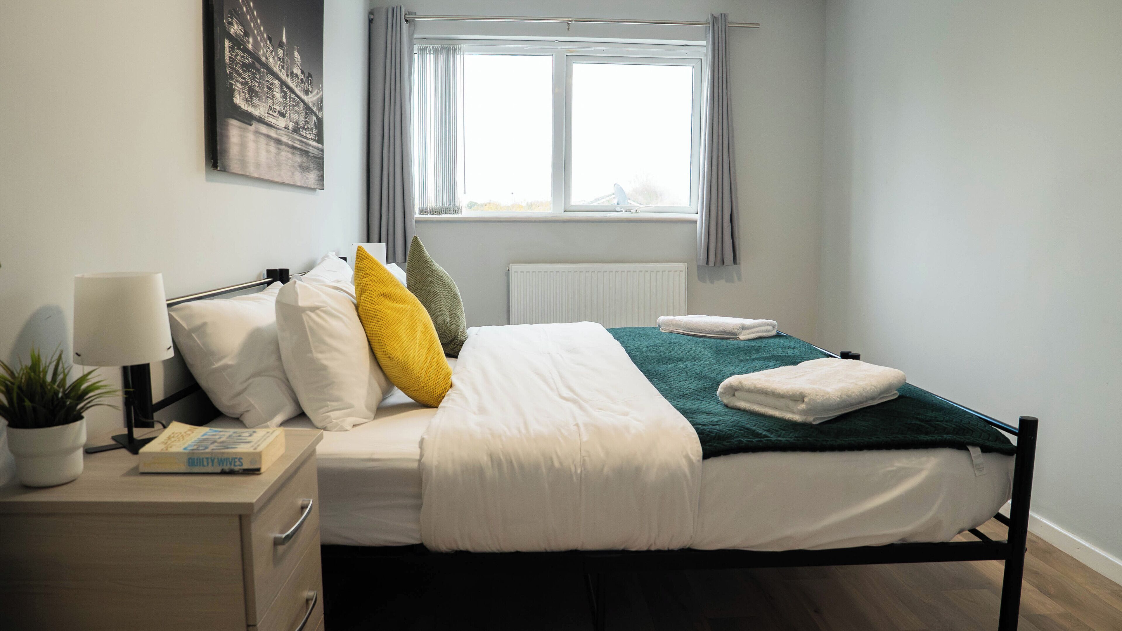 A neatly made bed with white and green bedding, a bedside lamp, a potted plant, and a framed picture on the wall.