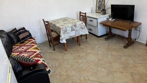 Un espacio habitable pequeño con un sofá, una pequeña mesa de comedor con mantel floreado, un escritorio de madera con televisor y un reloj de pared.