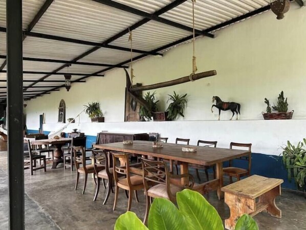 Un comedor con mesas y sillas de madera, techo alto con vigas de metal y plantas en macetas en la pared.
