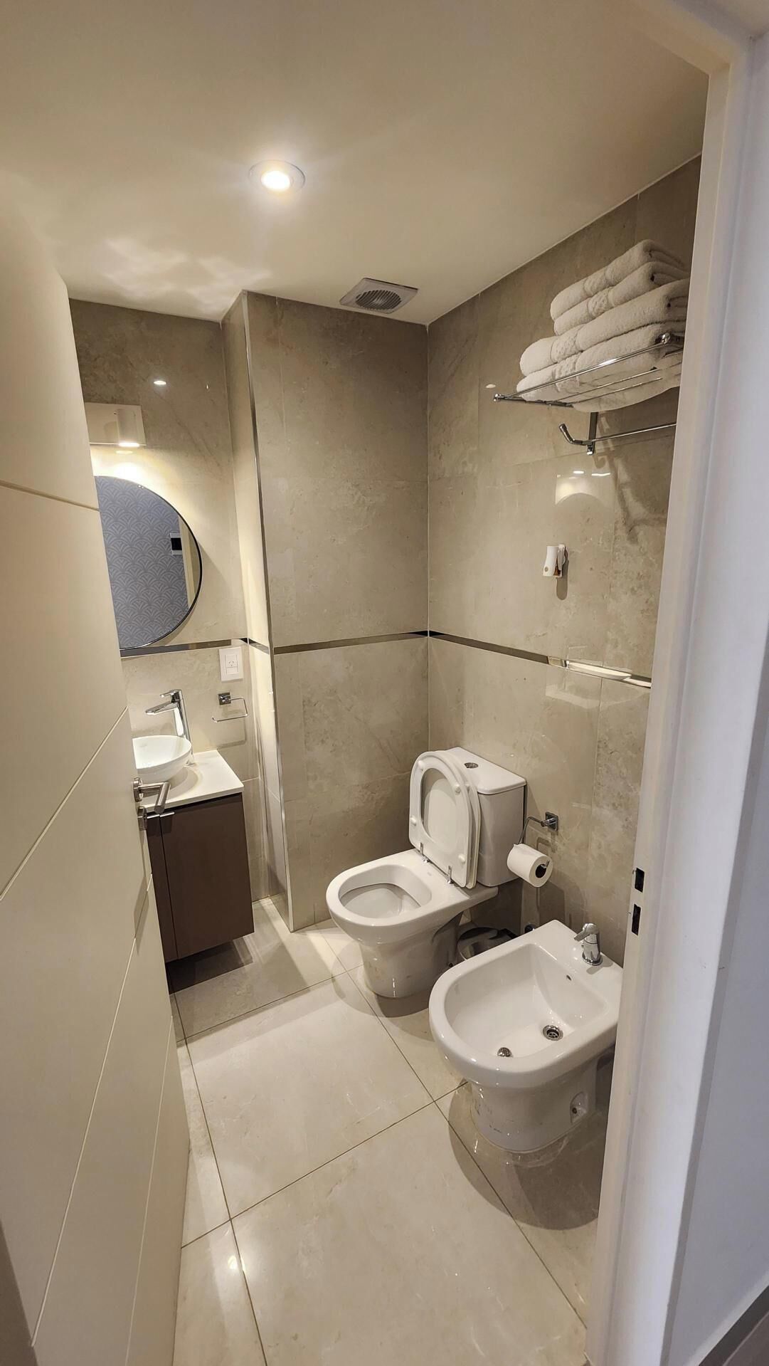 A modern bathroom with a toilet, bidet, and sink, and a towel rack with folded towels.