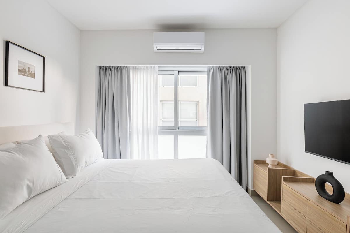 A hotel room with a large bed, a flat-screen TV, a wooden side table, and a window with curtains.