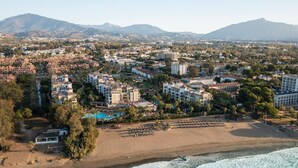 Vue aérienne d’une ville côtière avec une plage, des hôtels et une chaîne de montagnes en arrière-plan.