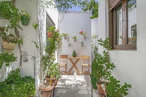 A narrow courtyard with a wooden table and chairs, surrounded by potted plants and a white wall.