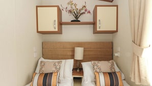 A hotel room with two beds, a wooden headboard, a nightstand with a lamp, and a small shelf with a flower arrangement. - Ty Seren (New Quay)