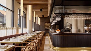 Un restaurante con mesas largas, sillas de mimbre, una zona de bar con mostrador y copas de vino colgando.