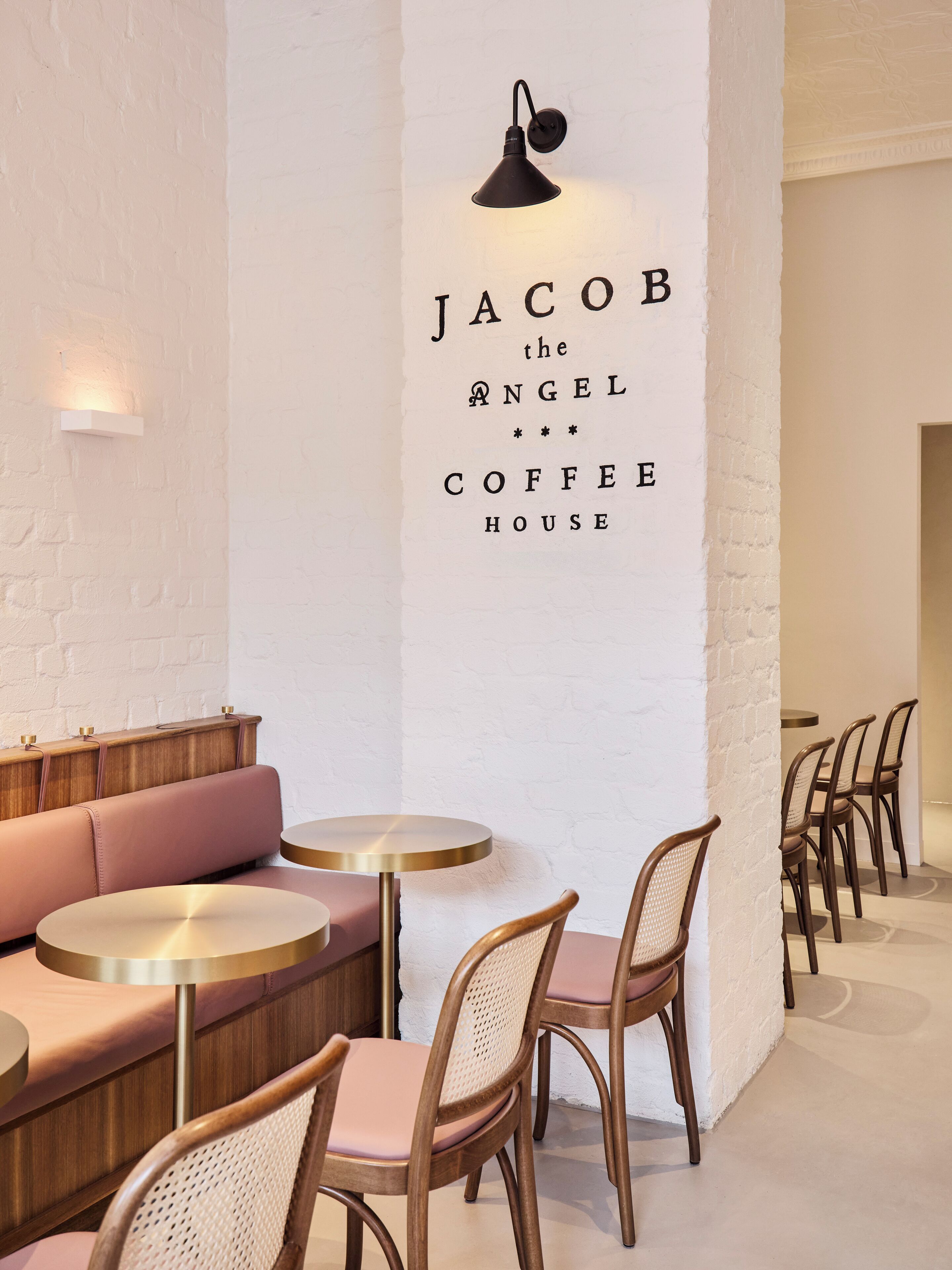 A cafe interior with a white brick wall, a black pendant light, and a sign reading 'Jacob the Angel Coffee House.' There are round tables and wooden chairs.