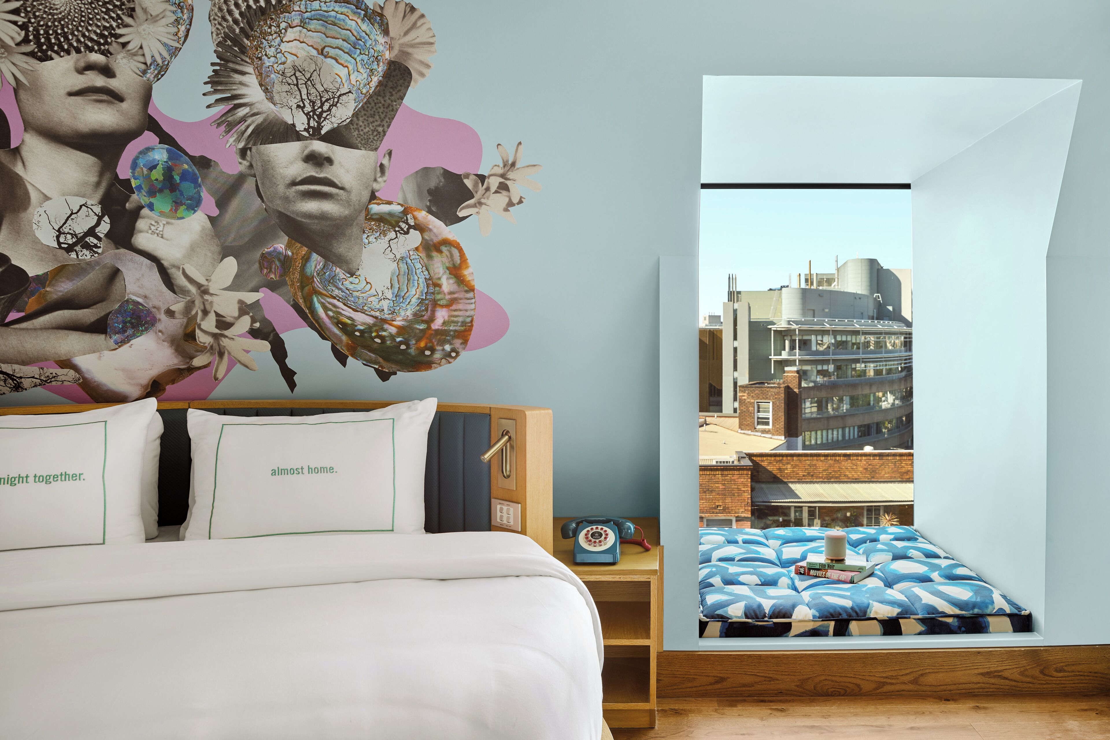Quarto com cama, almofadas, mesa de cabeceira, telefone e janela com vista para a cidade.