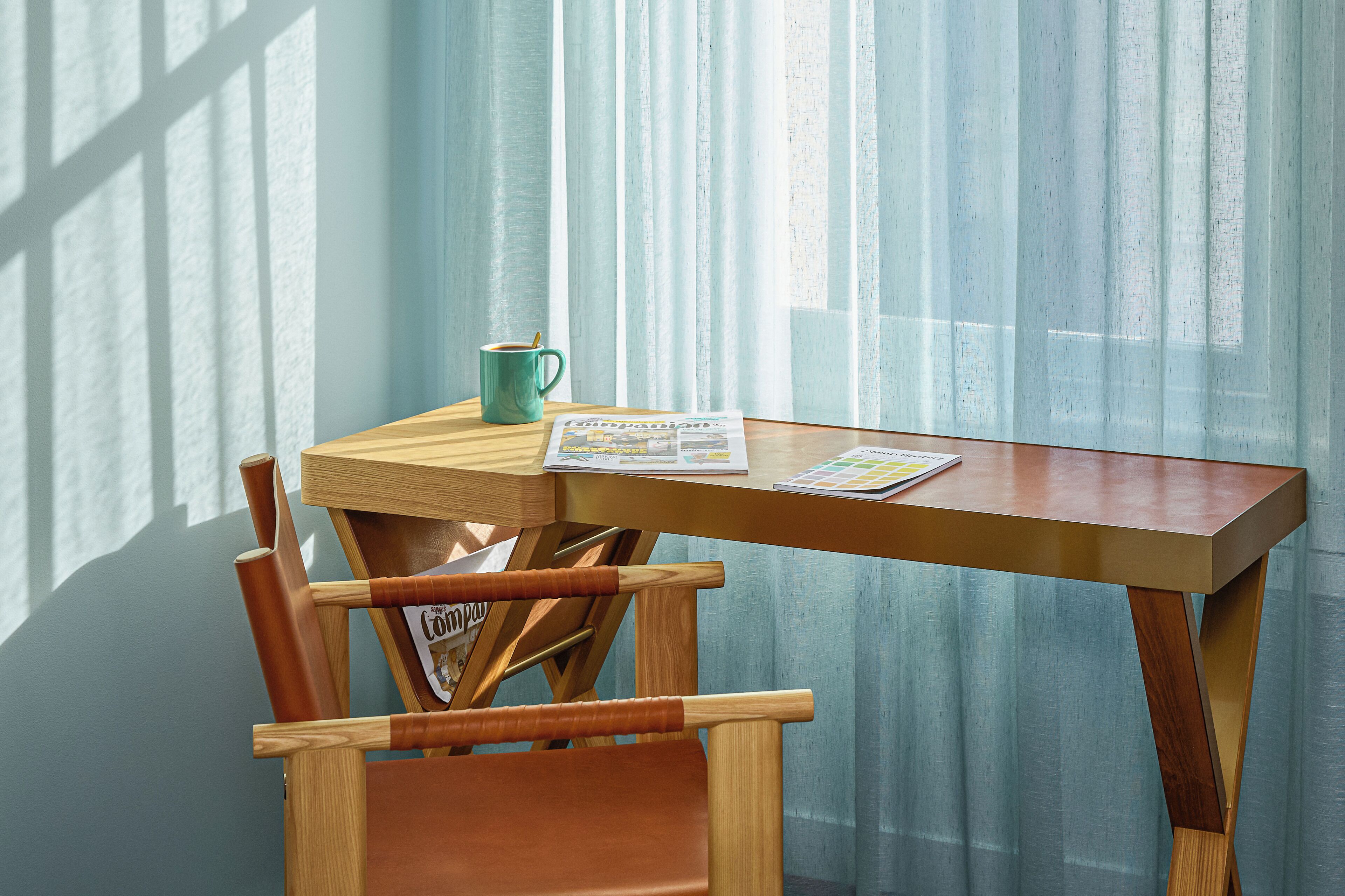 A wooden table with a magazine, a mug, and a chair in a room with light blue curtains.
