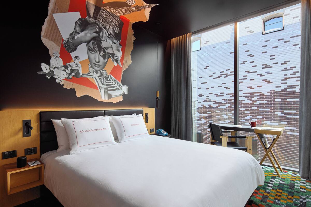 Een hotelkamer met een groot bed, een houten nachtkastje, een bureau met stoel, en een raam met uitzicht op een bakstenen gebouw.
