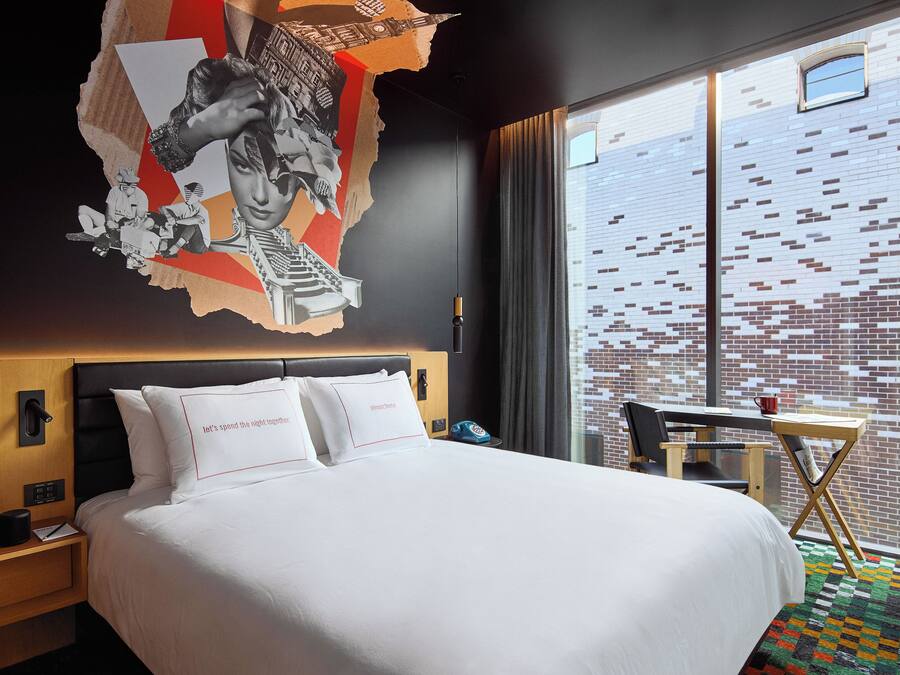 A hotel room with a large bed, a wooden nightstand, a desk with a chair, and a window with a view of a brick building.
