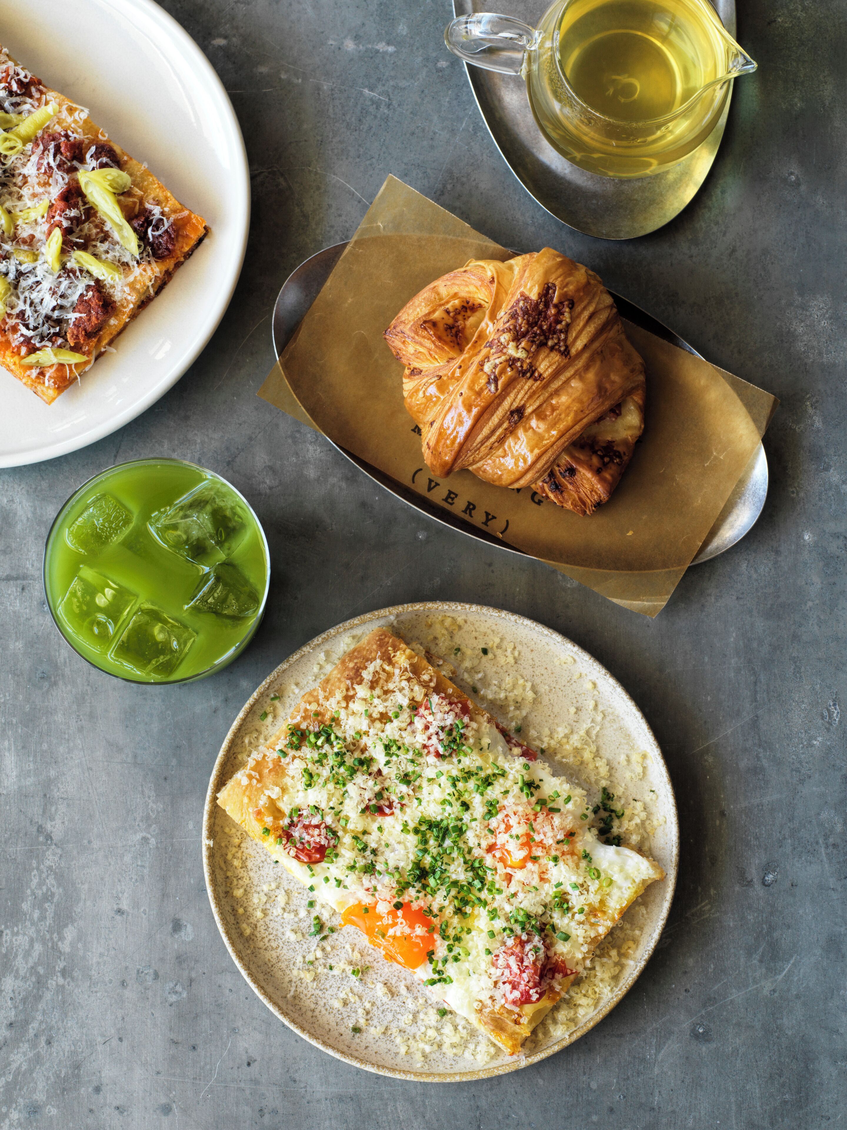 A meal with a croissant, a slice of pizza, and a plate of scrambled eggs with cheese and herbs, along with a green drink and a glass of wine on a table.