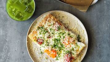 A meal with a croissant, a slice of pizza, and a plate of scrambled eggs with cheese and herbs, along with a green drink and a glass of wine on a table.