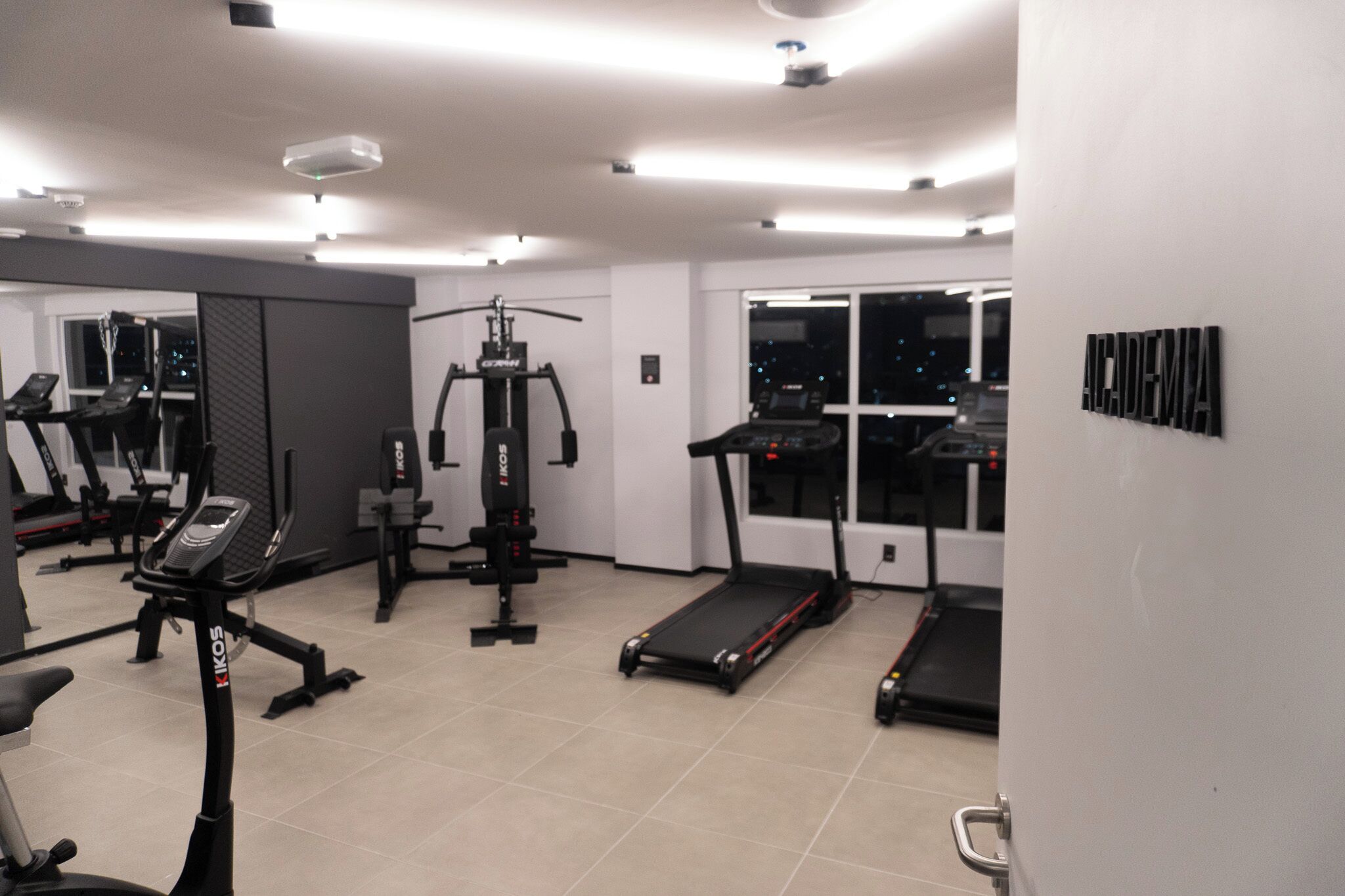A modern gym with various exercise equipment, including treadmills and weight machines, and large windows offering views of the city at night.