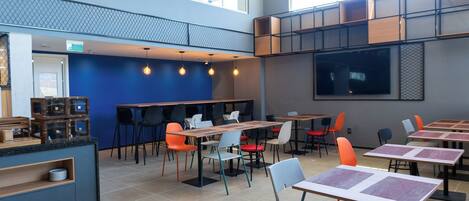 Una cafetería moderna con varias mesas y sillas, una barra con productos a la vista y grandes ventanales que dejan entrar luz natural.