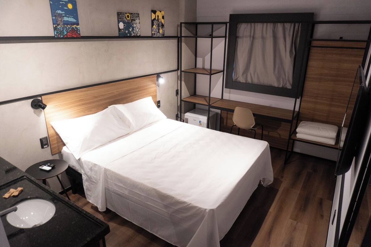 A hotel room with a bed, a desk, a chair, a small table, and a shelf with artwork.