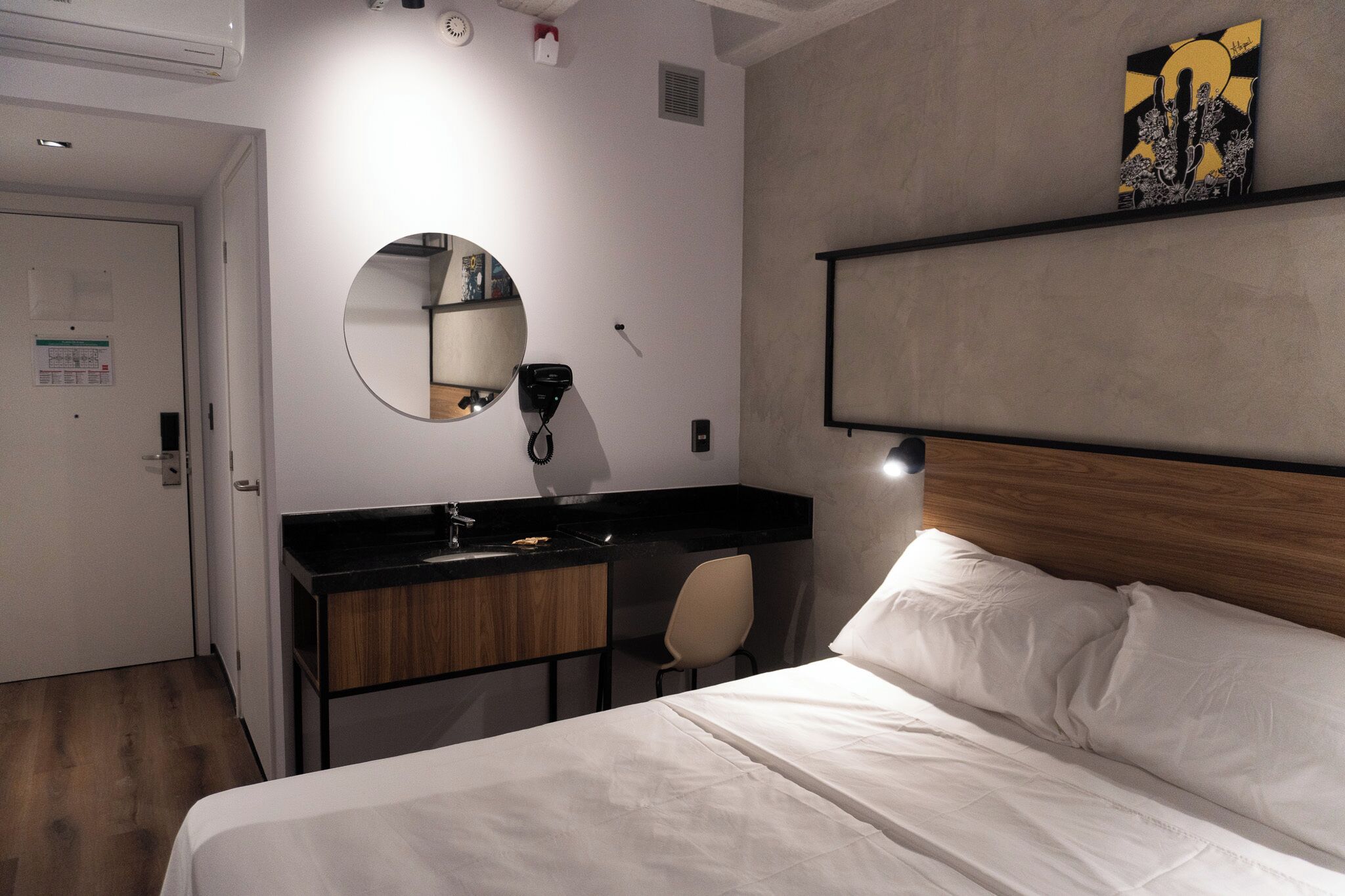 Une chambre d’hôtel avec un lit, un miroir rond, un plan de toilette noir avec un lavabo, un meuble en bois et une chaise.