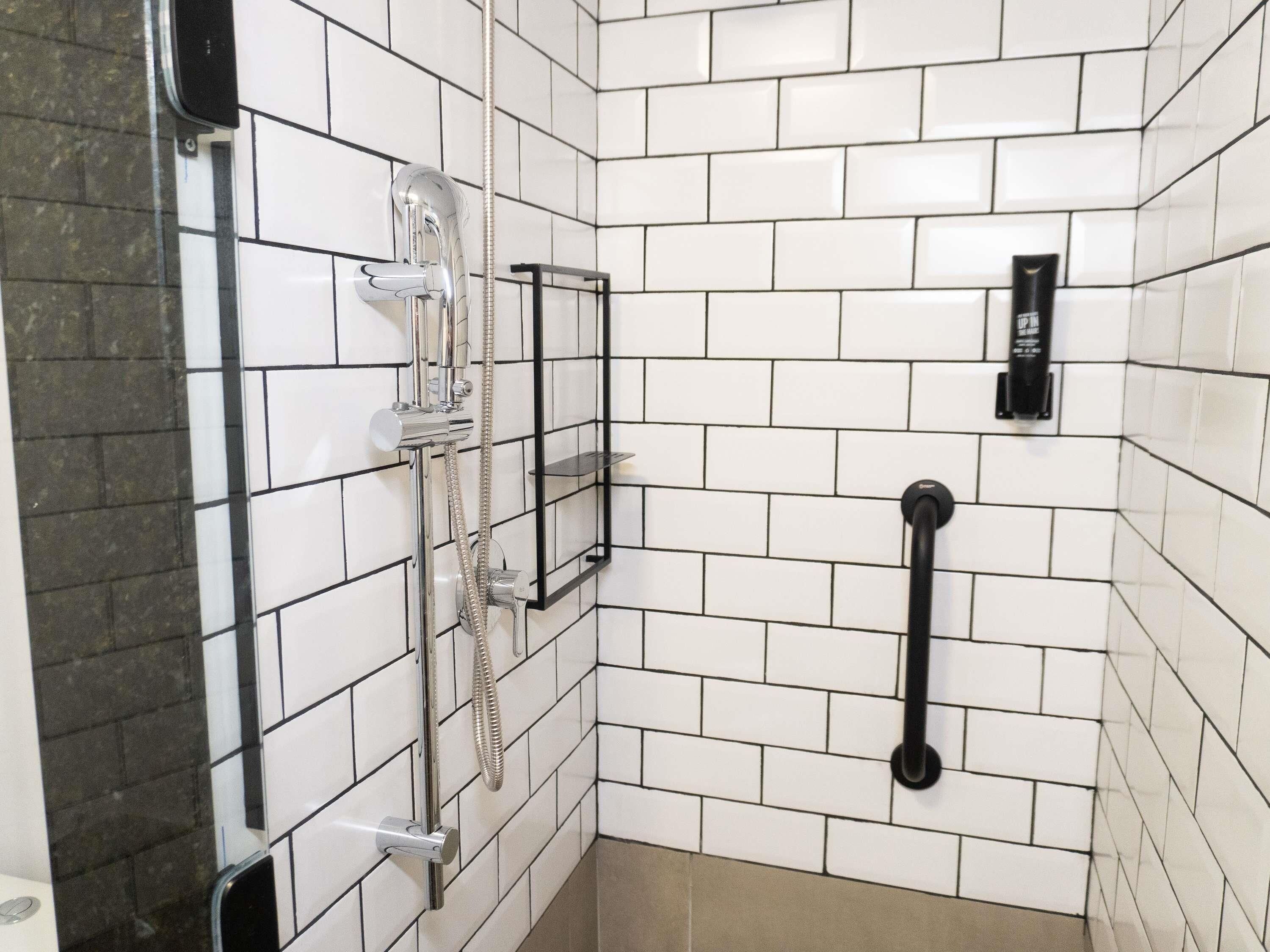 A bathroom with white tiled walls, a shower with a handheld showerhead, a black towel bar, and a soap dispenser.