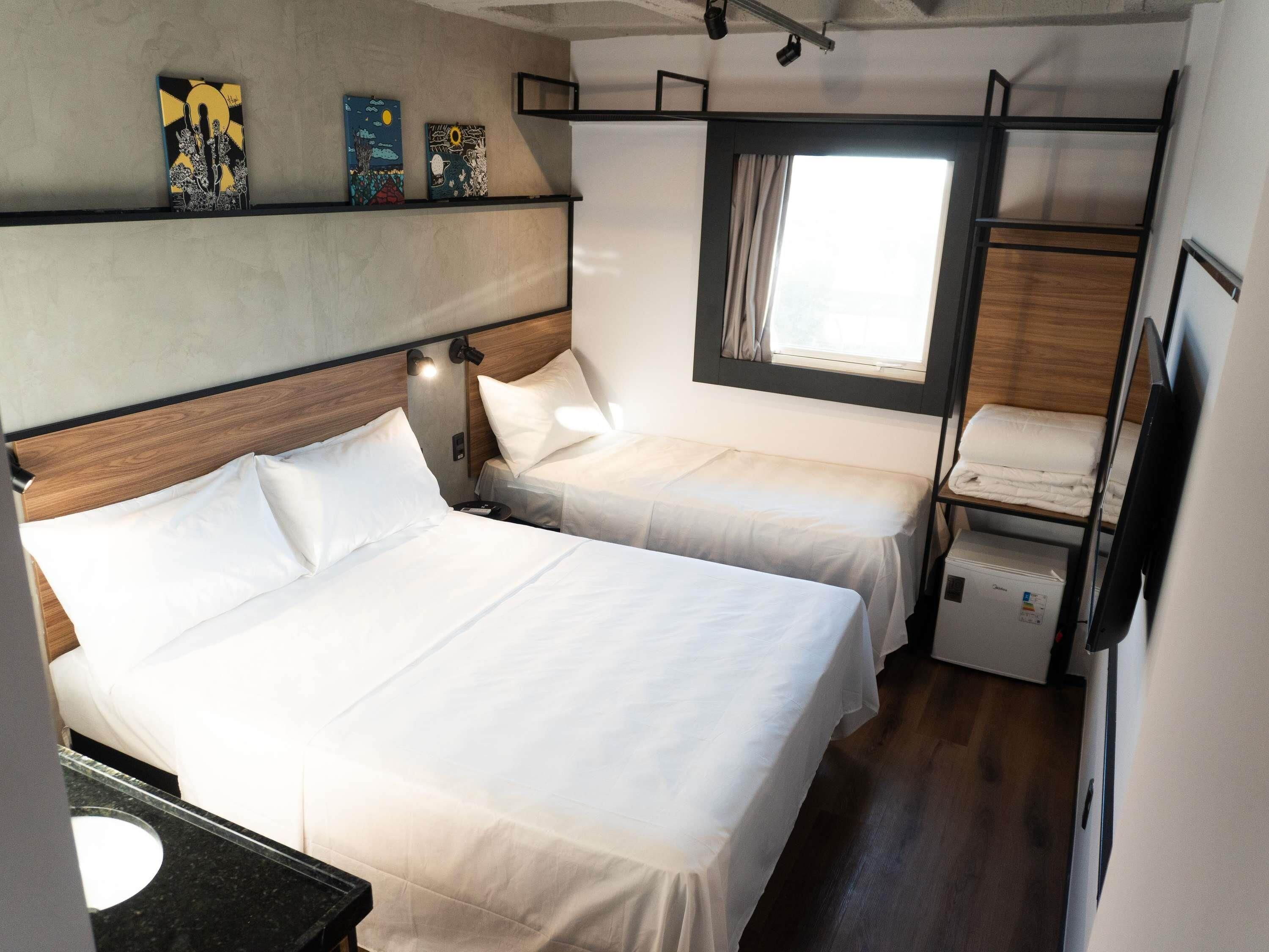 A compact bedroom with two beds, a window with curtains, and artwork on the wall.