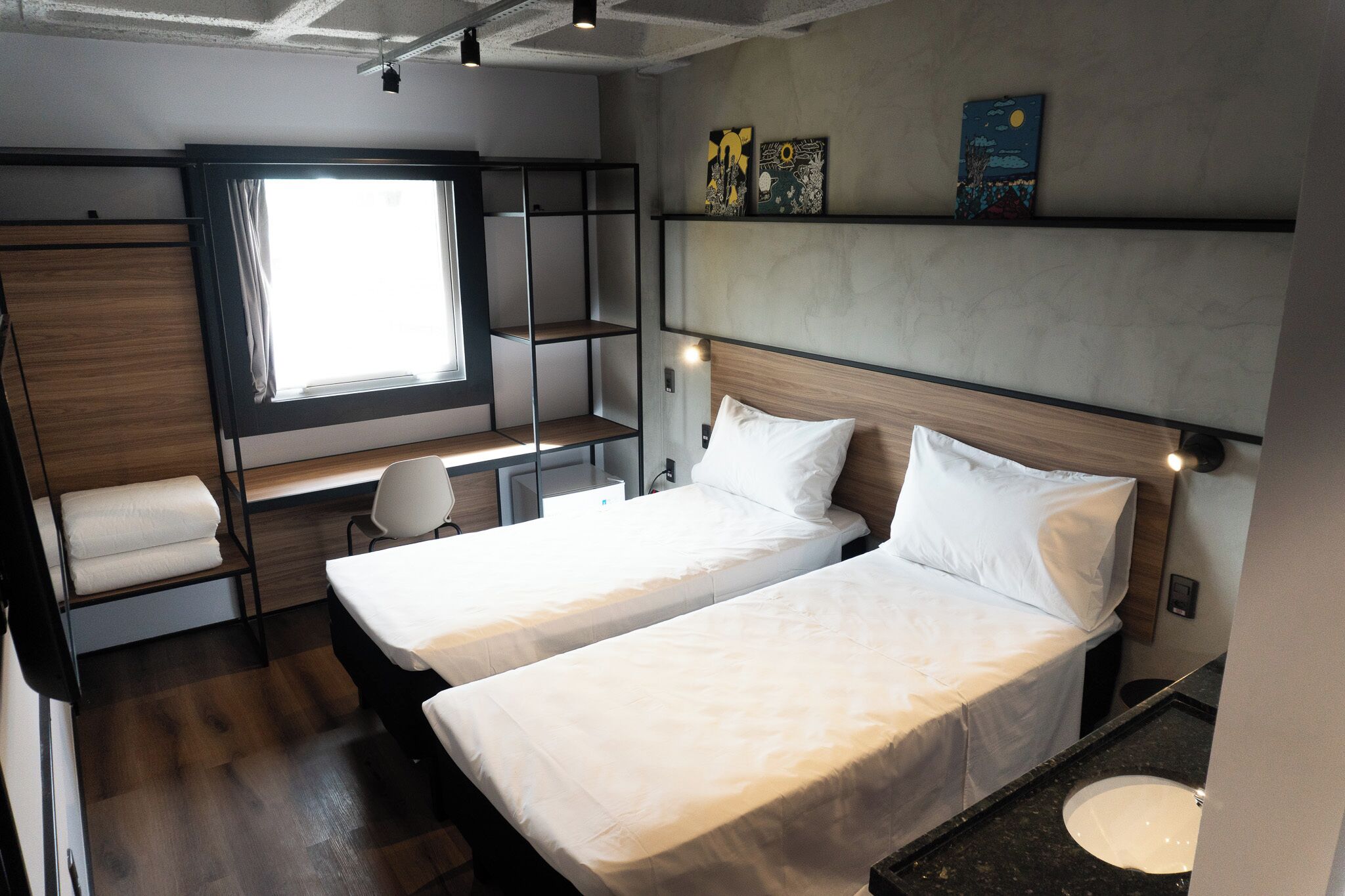 A compact hotel room with two single beds, a desk, a chair, a window with curtains, and artwork on the wall.