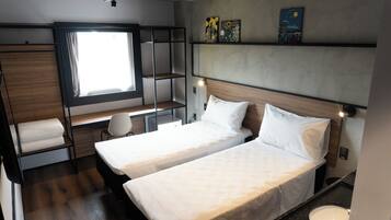 A compact hotel room with two single beds, a desk, a chair, a window with curtains, and artwork on the wall.